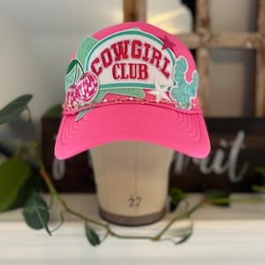 May include: A vibrant pink trucker cap with the text "COWGIRL CLUB" in white and green. The cap has a curved peak and is decorated with a cactus, stars, and cherries.