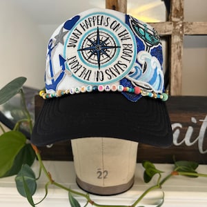 Puede incluir: Gorra de béisbol en blanco y negro con temática náutica. La gorra presenta parches con una rosa de los vientos, un ancla y un diseño de olas. El texto en la gorra dice "WHAT HAPPENS ON THE BOAT STAYS ON THE BOAT" y "LAKE BABE". Un adorno de cuentas está a lo largo del borde.