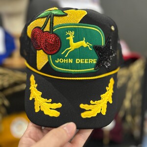 Może przedstawiać: Czarna czapka z daszkiem z zieloną naszywką z logo John Deere, żółtą gwiazdką z cekinów, dwiema czerwonymi wisienkami z cekinów, czarną gwiazdką z cekinów oraz dwoma żółtymi haftowanymi motywami liści na daszku z żółtym sznurkiem.