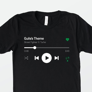 May include: Black t-shirt featuring a white music player graphic. The graphic displays the song "Guile's Theme" from Street Fighter II: Turbo playing. The music player has a play button, rewind button, fast forward button, and a repeat button.