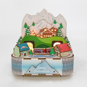 May include: A wooden, illuminated winter village scene with a mountain backdrop. The scene includes houses, trees, a train, and the text "Snow Paradise". The houses have red, blue, and tan roofs. The train is red and the trees are green.