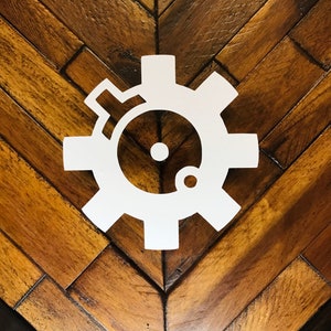 May include: A white gear-shaped design with a circular center and a smaller circle, set against a wooden herringbone pattern background. The gear has multiple teeth and a detailed inner design.