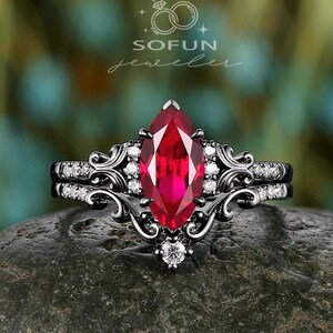 May include: A marquise-cut ruby engagement ring with a black metal band. The ring features a central red gemstone, surrounded by smaller white stones and ornate filigree detailing. The ring is set on a dark gray stone.