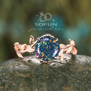 May include: A rose gold ring with a leafy design and a round, iridescent opal gemstone. The ring is set on a gray rock.