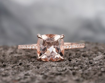 May include: A rose gold engagement ring with a cushion-cut morganite center stone and a diamond halo. The ring is set on a delicate band with pave diamonds.