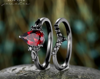 May include: Two-piece ring set featuring a pear-shaped, deep red gemstone accented with black stones. The rings have a dark, gunmetal finish. The set is displayed on a textured, gray stone surface.