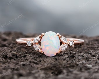 May include: A rose gold ring featuring a central oval-shaped iridescent opal stone. The setting is accented on each side by two marquise-cut clear stones and one small round clear stone, creating a symmetrical arrangement along the thin metal band.