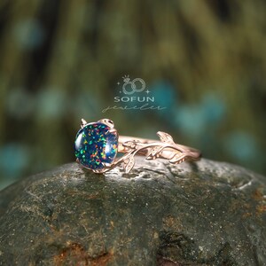 May include: A rose gold ring with a green and black opal gemstone set in a delicate branch design. The ring is on a dark green rock. SOFUN jeweler.