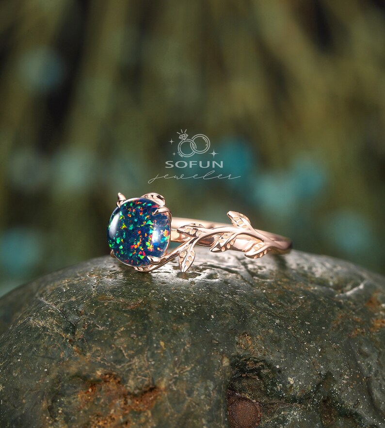 May include: A rose gold ring with a green and black opal gemstone set in a delicate branch design. The ring is on a dark green rock. SOFUN jeweler.