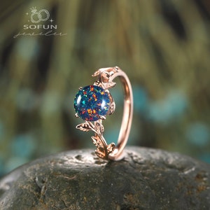 May include: A rose gold ring with a leafy design and a round, iridescent opal gemstone. The ring is set on a gray rock.
