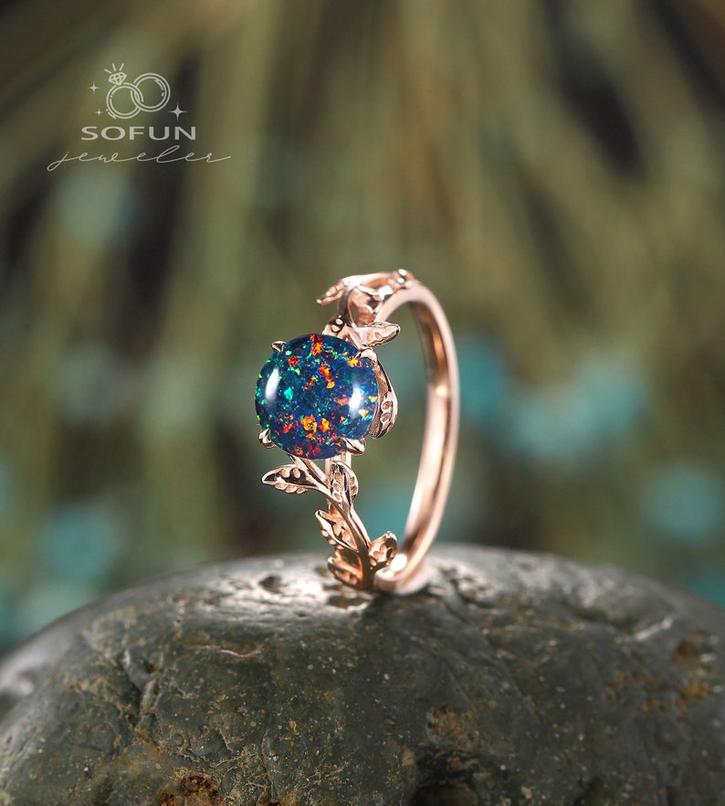 May include: A rose gold ring with a leafy design and a round, iridescent opal gemstone. The ring is set on a gray rock.