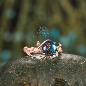 May include: A rose gold ring with a green and red opal gemstone set in a delicate branch design. The ring is on a gray rock.