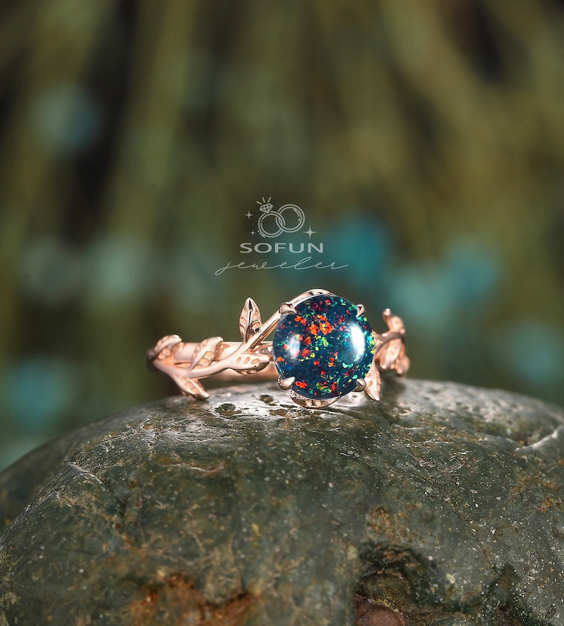 May include: A rose gold ring with a green and red opal gemstone set in a delicate branch design. The ring is on a gray rock.