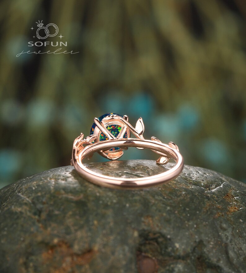 May include: A rose gold ring with a large oval opal gemstone set in a delicate branch design. The ring is on a gray rock.