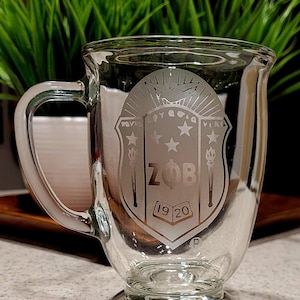 May include: A clear glass mug with a handle, featuring an etched design of a shield with the letters "ZOB" and the numbers "1920". The shield also includes a dove, three stars, and two torches.