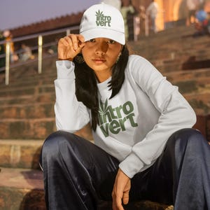 Könnte beinhalten: Weißes Baseball-Cap und langärmliges Sweatshirt mit dem grünen Text "Intro Vert" und einem Cannabisblatt-Design. Das Logo von The Gas Station ist sichtbar. Die Person trägt eine dunkelblaue Hose.