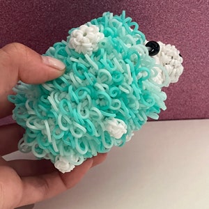May include: A handmade, fluffy, blue and white toy animal, possibly a sheep or dog, made from looped rubber bands. The toy has small white flower-like accents and black bead eyes. The toy is held in a hand.