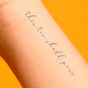 May include: A temporary tattoo with the text "this too shall pass" written in cursive.