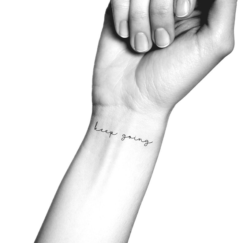 Pode incluir: Uma fotografia a preto e branco de um pulso com uma tatuagem simples que diz "keep going" em cursivo.