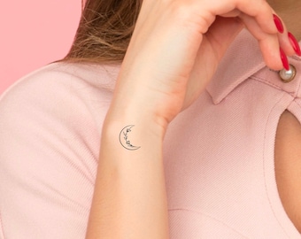 May include: A simple black line drawing of a crescent moon with a floral wreath around it. The tattoo is on a person's wrist.