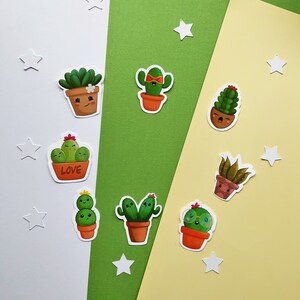 May include: A collection of colorful cactus stickers with smiling faces and various designs. The stickers are in terracotta pots and feature details like flowers and bow ties. White star-shaped cutouts are scattered around the stickers.