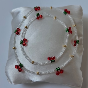 May include: A white beaded necklace with red and green beads in the shape of cherries. The necklace is layered and has gold beads interspersed throughout.