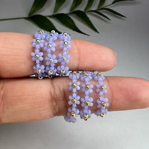 May include: Set of four beaded rings with a flower design. The rings are made with light purple and gold beads.