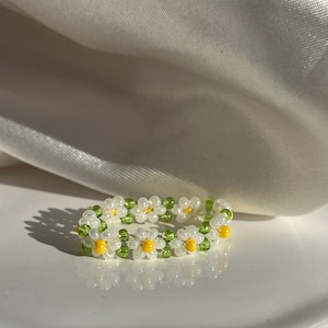 May include: A white and yellow daisy flower ring made with small beads. The ring is on a white surface.