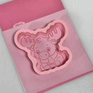 May include: A light pink plastic cookie cutter featuring a cartoon moose design with large antlers and a scarf. The cutter is attached to a pink backing card that displays the text S4U C with a small heart symbol between the letters.