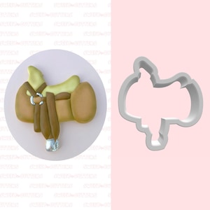 May include: A cookie cutter and a decorated cookie in the shape of a western saddle. The cookie is tan, brown, and green with silver accents. The cookie cutter is white plastic. The background is pink and white.
