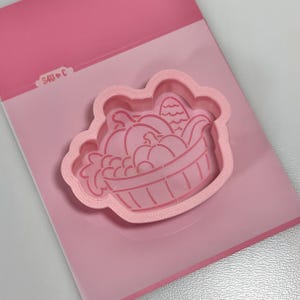 May include: A light pink plastic cookie cutter featuring a detailed design of a harvest basket filled with pumpkins and corn. The cutter has a raised edge and includes the text S4U C embossed on the top left corner of the frame.