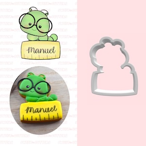 May include: A green cartoon caterpillar wearing glasses, holding a yellow ruler with the name "Manuel" written on it. The image also shows a cookie version of the caterpillar and a cookie cutter.