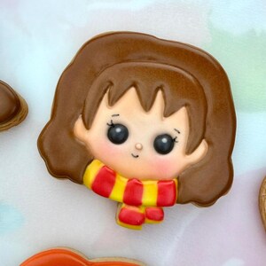 May include: A sugar biscuit shaped like a cartoon character head with brown hair, large black eyes, and a small nose. The character wears a scarf with alternating horizontal red and yellow stripes. The icing is smooth with a glossy finish.