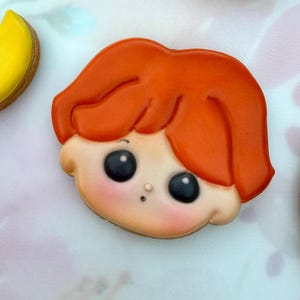 May include: A sugar biscuit shaped like a cartoon face with bright orange hair, large black eyes with white highlights, a small nose, and a tiny mouth. The skin tone is a light peach colour with soft pink blush on the cheeks, featuring smooth icing.