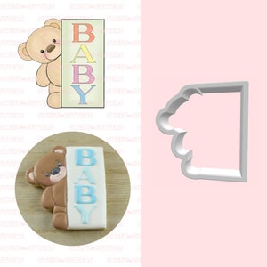 Op de afbeelding: Een koekjesvormpje in de vorm van een teddybeer die achter een bordje met de tekst "BABY" in kleurrijke letters kijkt. De koekjesvormpje is gemaakt van wit plastic.