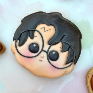 May include: A decorated sugar cookie shaped like a cartoon face with black hair, large round black glasses, and a small lightning bolt symbol on the forehead. The cookie features a light skin tone, rosy cheeks, and a simple, minimalist facial expression.