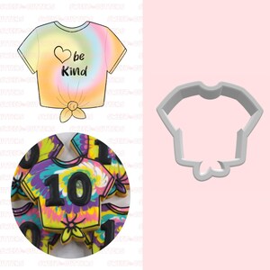 May include: A cookie cutter in the shape of a tie-dye t-shirt with a bow at the bottom. The cutter is made of gray plastic and is shown on a pink background. The image also shows a close-up of a tie-dye cookie decorated with black icing.