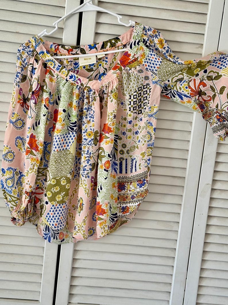 Puede incluir: Una blusa de manga larga con base rosa claro y un patr&oacute;n de patchwork multicolor con dise&ntilde;os florales, de cachemira y geom&eacute;tricos. La prenda tiene un escote en V, tela fruncida en los hombros y pu&ntilde;os el&aacute;sticos en las mangas.