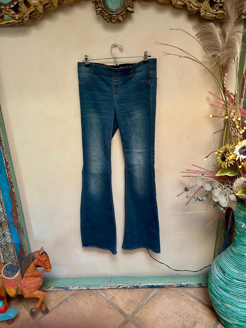 Puede incluir: Vaqueros acampanados de mezclilla azul colgados de una percha de metal. Los vaqueros tienen cintura alta y un lavado descolorido. Las perneras se ensanchan desde la rodilla. Los vaqueros se exhiben contra una pared neutra.