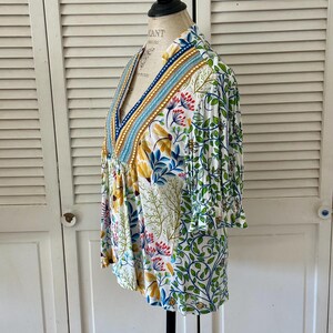 May include: A white, floral-print top with a V-neck and three-quarter sleeves. The top features a colorful botanical pattern with yellow, blue, and red flowers, and green leaves. The neckline is trimmed with blue and gold accents.