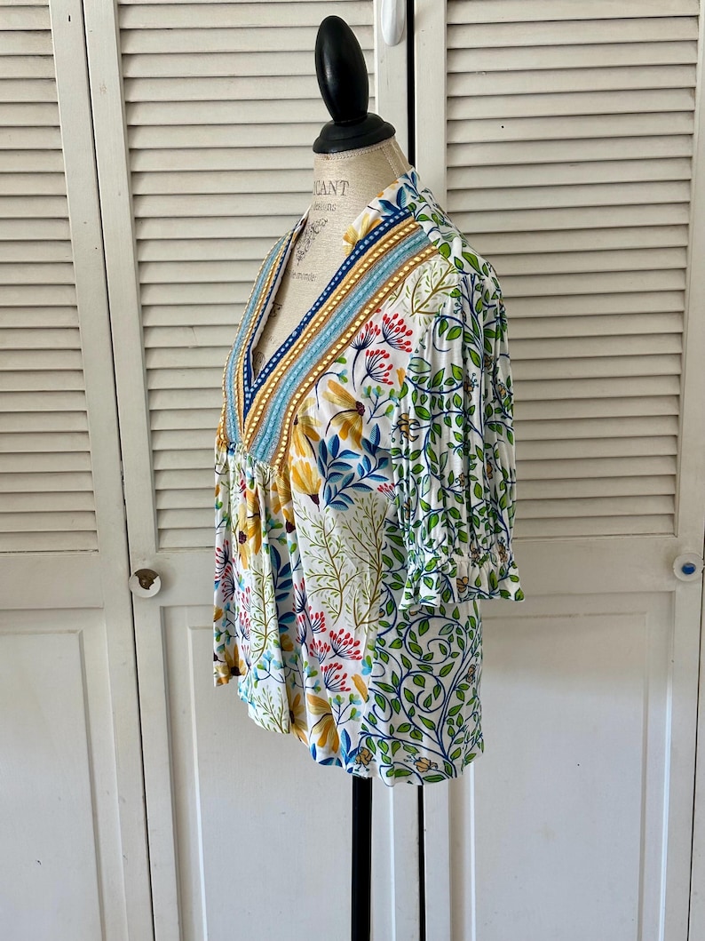 May include: A white, floral-print top with a V-neck and three-quarter sleeves. The top features a colorful botanical pattern with yellow, blue, and red flowers, and green leaves. The neckline is trimmed with blue and gold accents.