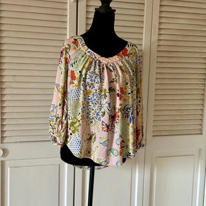 Puede incluir: Una blusa de manga larga con una base de color rosa claro y un estampado floral y de cachemira multicolor. La prenda presenta un escote en pico, hombros fruncidos y pu&ntilde;os el&aacute;sticos en las mangas, creando una silueta holgada y fluida.