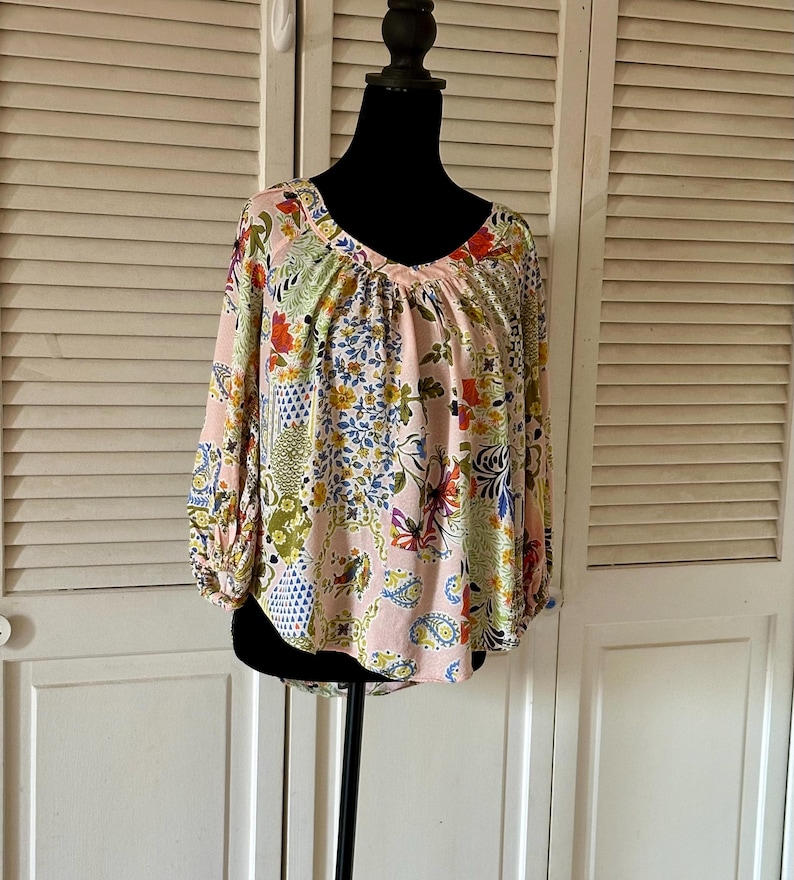 Puede incluir: Una blusa de manga larga con una base de color rosa claro y un estampado floral y de cachemira multicolor. La prenda presenta un escote en pico, hombros fruncidos y pu&ntilde;os el&aacute;sticos en las mangas, creando una silueta holgada y fluida.