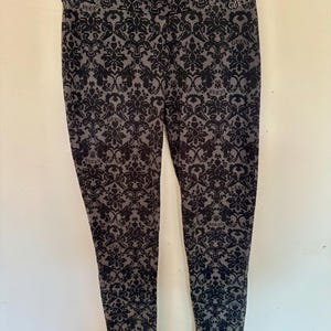 May include: Black and gray patterned leggings with a floral design. The leggings have the word "GUESS" printed on the waistband. The leggings are hanging on a hanger against a white wall.