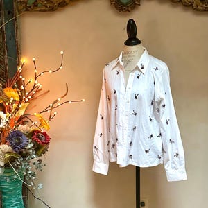 May include: White button-down shirt with long sleeves, adorned with small, embroidered black and burgundy figures. The shirt is displayed on a mannequin, showcasing its tailored fit and design. A classic, versatile garment.