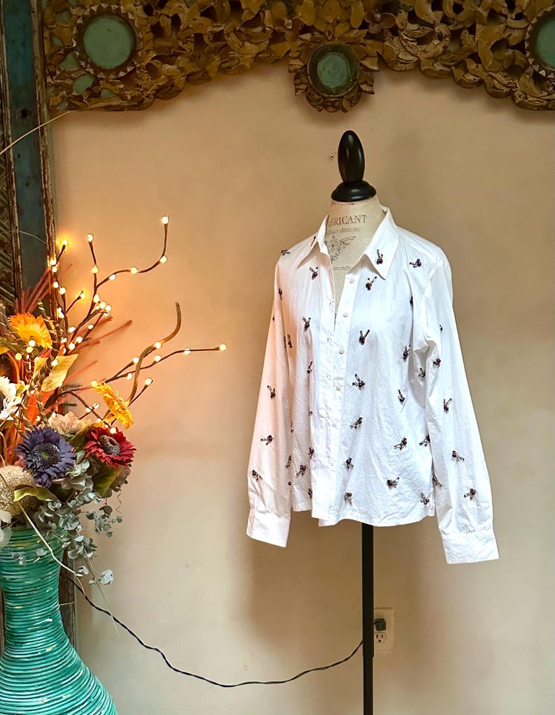May include: White button-down shirt with long sleeves, adorned with small, embroidered black and burgundy figures. The shirt is displayed on a mannequin, showcasing its tailored fit and design. A classic, versatile garment.