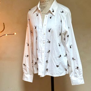 May include: White button-down shirt with long sleeves, adorned with small, dark-colored decorative elements. The shirt is displayed on a mannequin, showcasing its tailored fit and classic design. The shirt has a collared neckline.