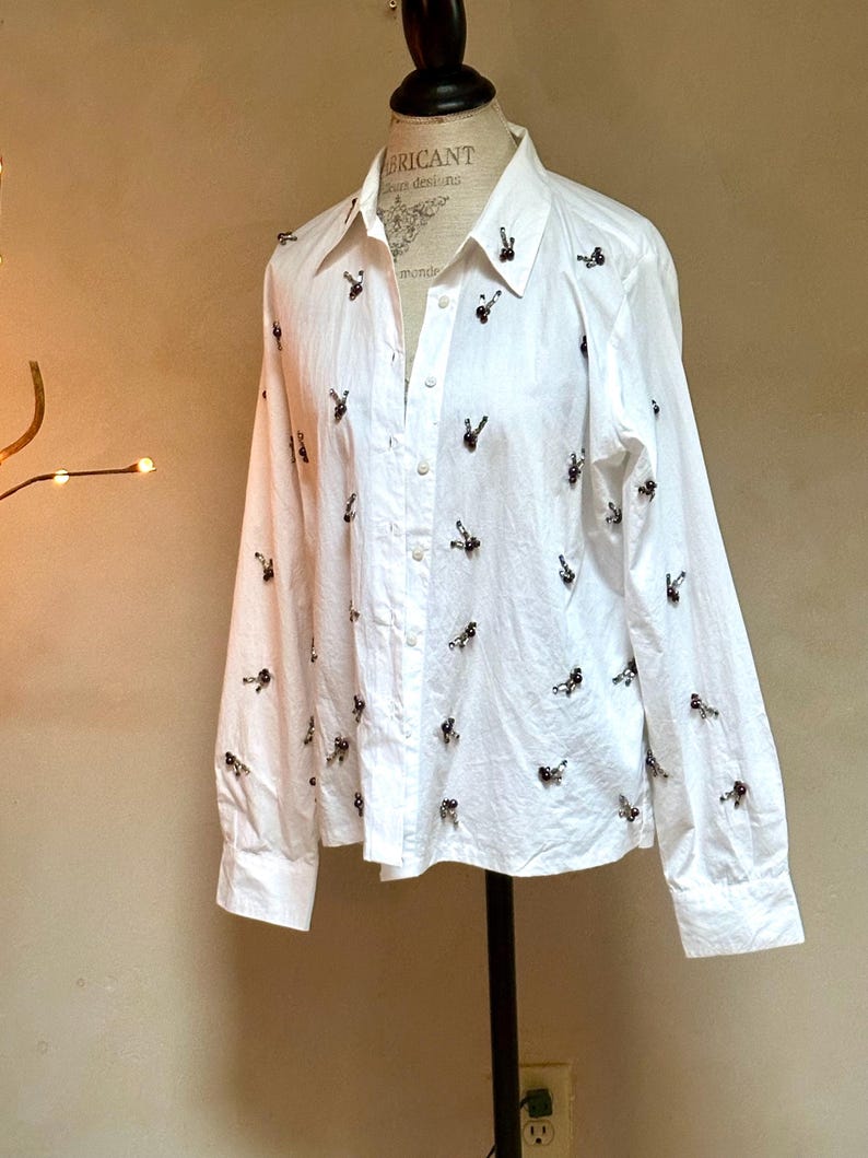 May include: White button-down shirt with long sleeves, adorned with small, dark-colored decorative elements. The shirt is displayed on a mannequin, showcasing its tailored fit and classic design. The shirt has a collared neckline.
