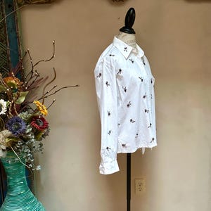 May include: White button-down shirt with long sleeves, adorned with small, embroidered bee motifs. The shirt is displayed on a mannequin, showcasing its tailored fit and classic design. The collar is neatly folded, and the cuffs are buttoned.