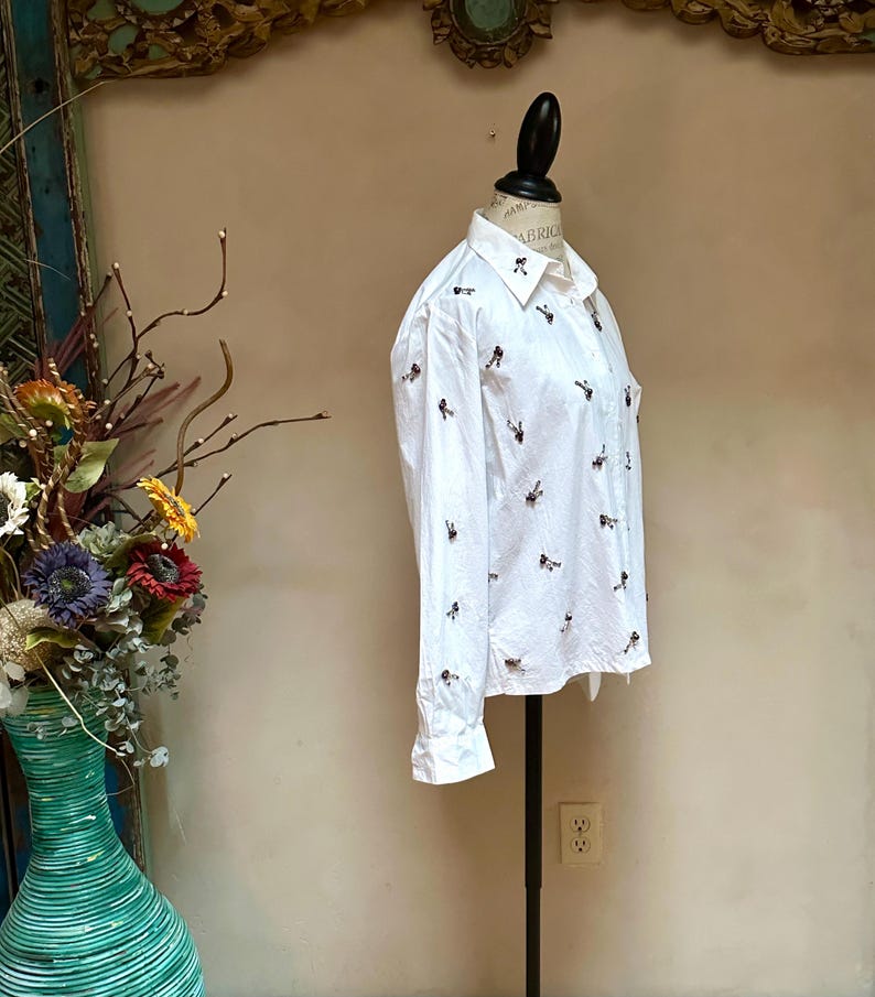 May include: White button-down shirt with long sleeves, adorned with small, embroidered bee motifs. The shirt is displayed on a mannequin, showcasing its tailored fit and classic design. The collar is neatly folded, and the cuffs are buttoned.